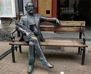 Imagem de uma estátua de Adolphe Sax, inventor do saxofone, sentado num banco de praça segurando o instrumento, localizada na Bélgica, seu país de origem.