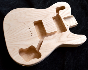 Imagem do corpo maciço de uma guitarra sem acabamento mostrando os espaços de encaixe dos captadores, knobs e chave-seletora onde se encontra a parte elétrica do instrumento.